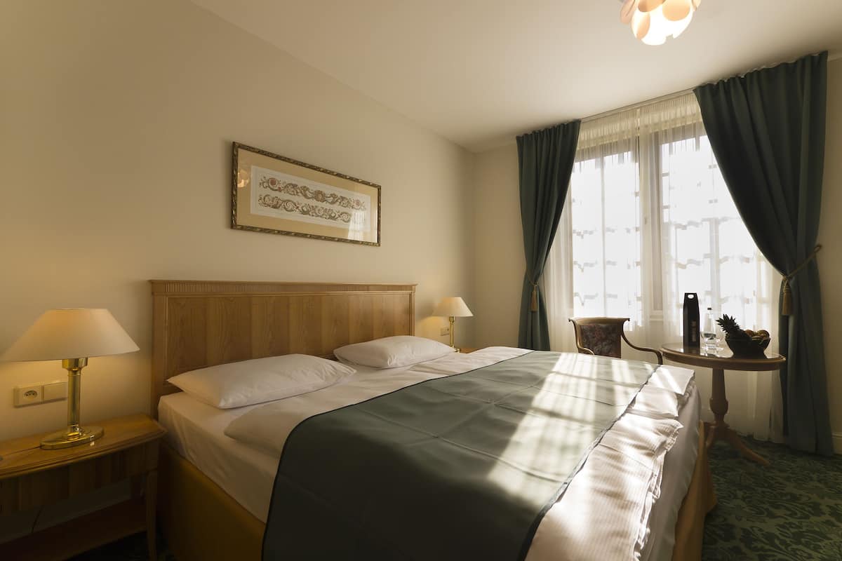 Een hotelkamer met een groot bed, twee bedlampjes, een fotokader aan de muur, een klein tafeltje met een lamp en een raam met gordijnen.