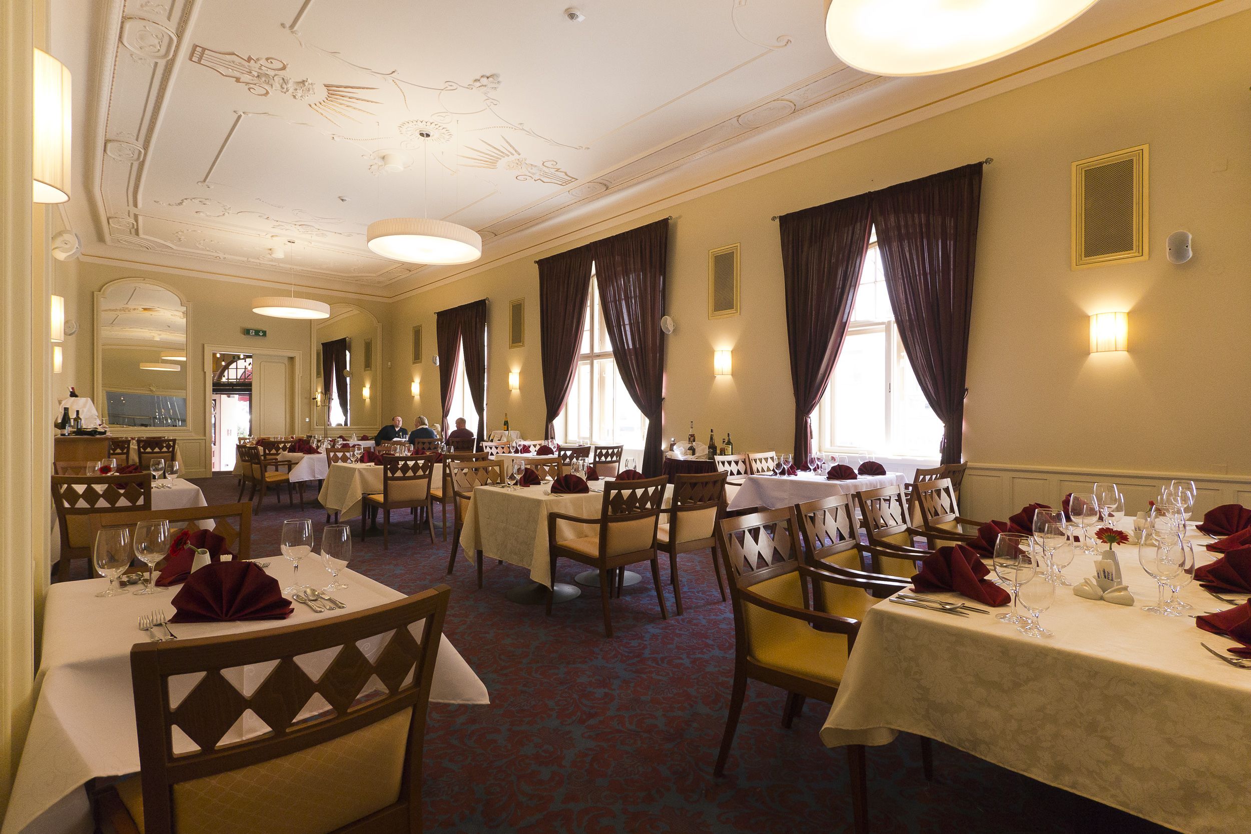 A dining area with tables set for a meal, featuring chandeliers, curtains, and wall-mounted lights.