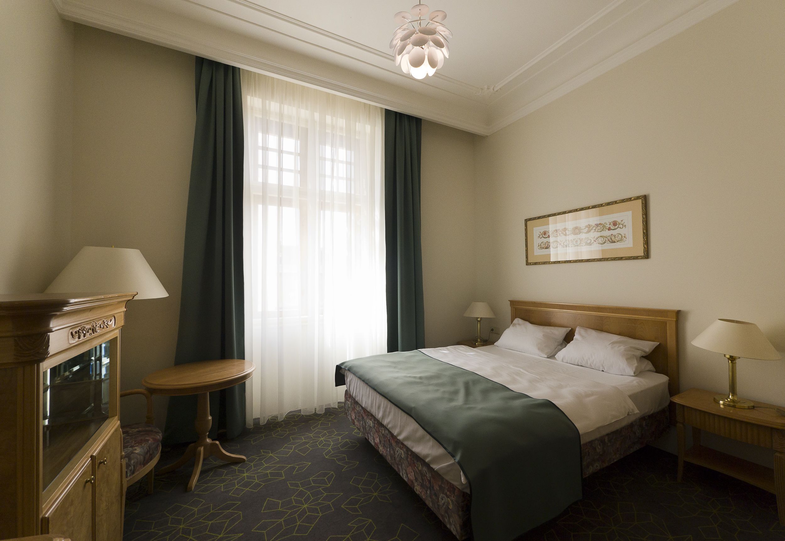 Una habitación de hotel con una cama grande, dos mesitas de noche con lámparas, un mueble de madera, una pequeña mesa redonda y un amplio ventanal con cortinas.