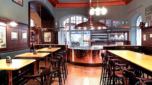 Interior de un restaurante con mesas y sillas de madera, una barra de cobre y un amplio ventanal de arco.