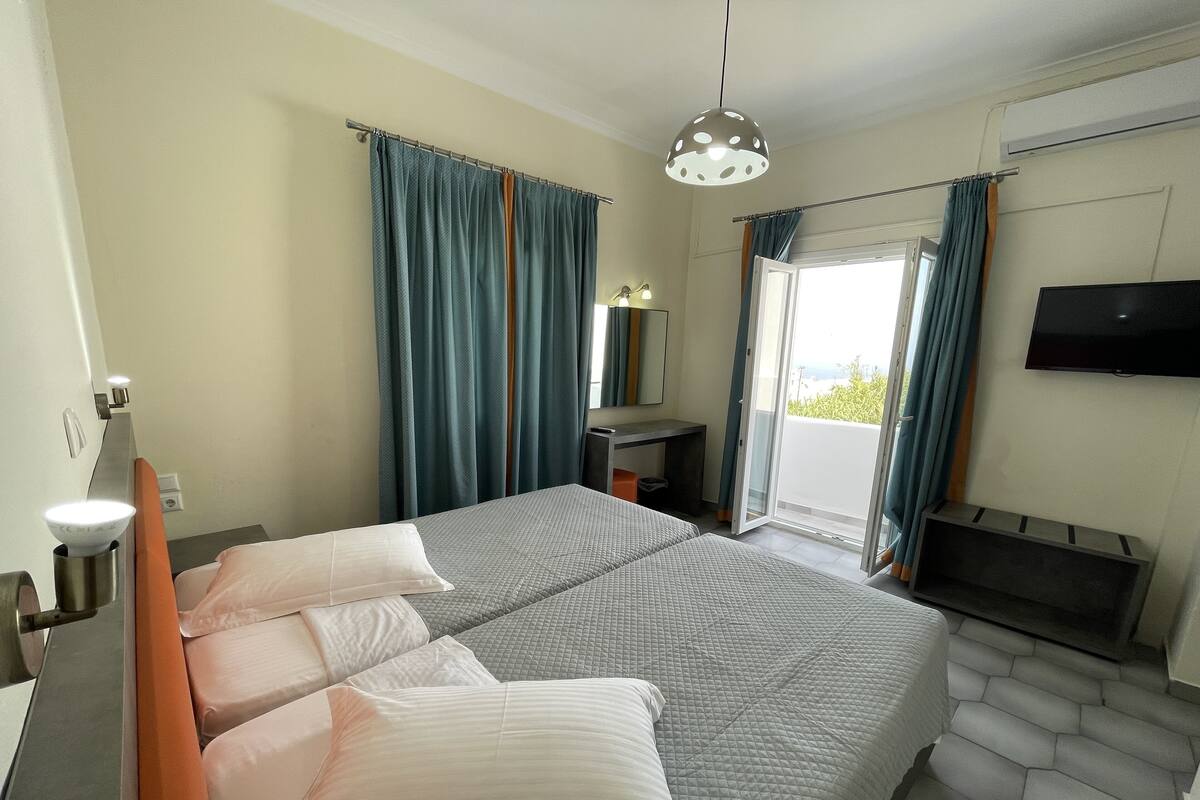 Habitación de hotel con cama, escritorio, televisor y balcón con cortinas.