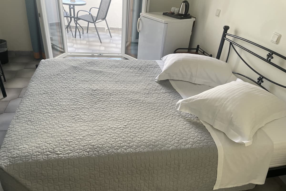 A bed with a quilted cover, a bedside table with a kettle, and a chair in a room with a tiled floor.