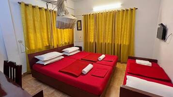 Een hotelkamer met twee bedden, elk voorzien van rode beddengoed en witte kussens. Er is een plafondventilator, een luchtbevochtiger aan de muur en een klein tafeltje met een stoel.