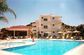 Un área de piscina con sillones de descanso y sombrillas, un edificio con varios balcones y una palmera.