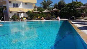 A swimming pool with clear blue water, surrounded by lounge chairs and umbrellas, with a building in the background. - Summerdays Studios (Rhodes)