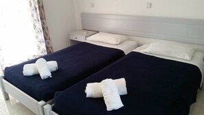 A hotel room with two beds, each with a dark blue bedspread and white pillows. There is a nightstand with a lamp between the beds. - Summerdays Studios (Rhodes)