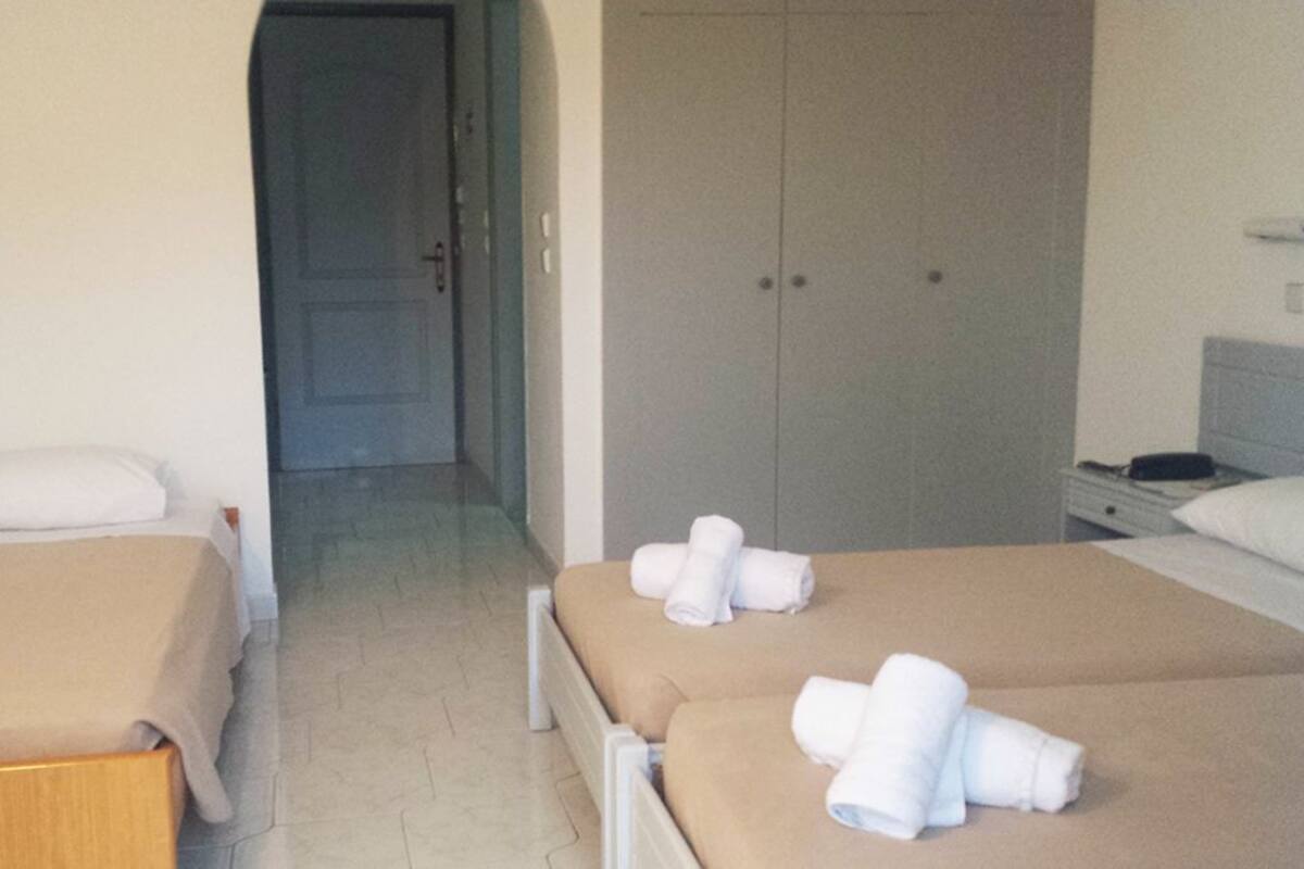 A hotel room with two beds, each with a beige bedspread and white pillows. There is a door leading to a hallway and a wardrobe in the background.