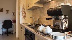 Una cocina con estufa, lavamanos y cafetera. Hay una silla y una mesa con platos y tazas.