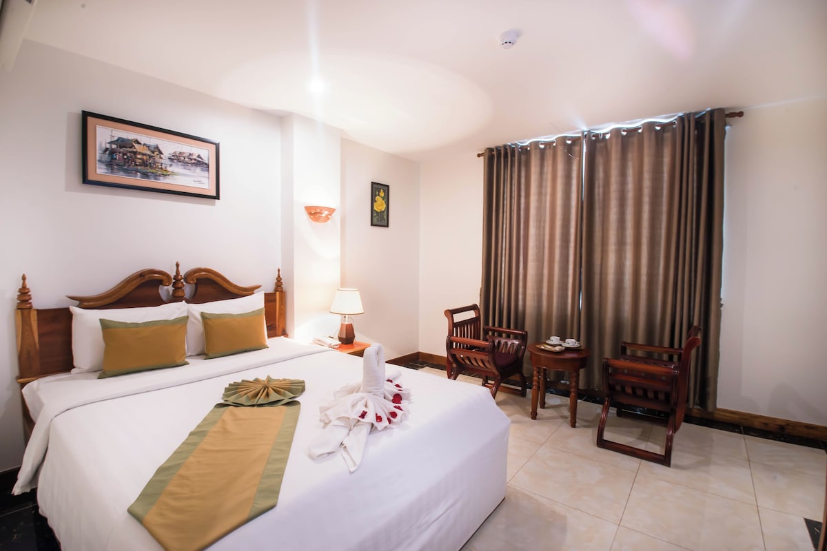 Quarto de hotel com uma cama grande, mesa de cabeceira com abajur, cadeira, pequena mesa com xícaras e cortina.