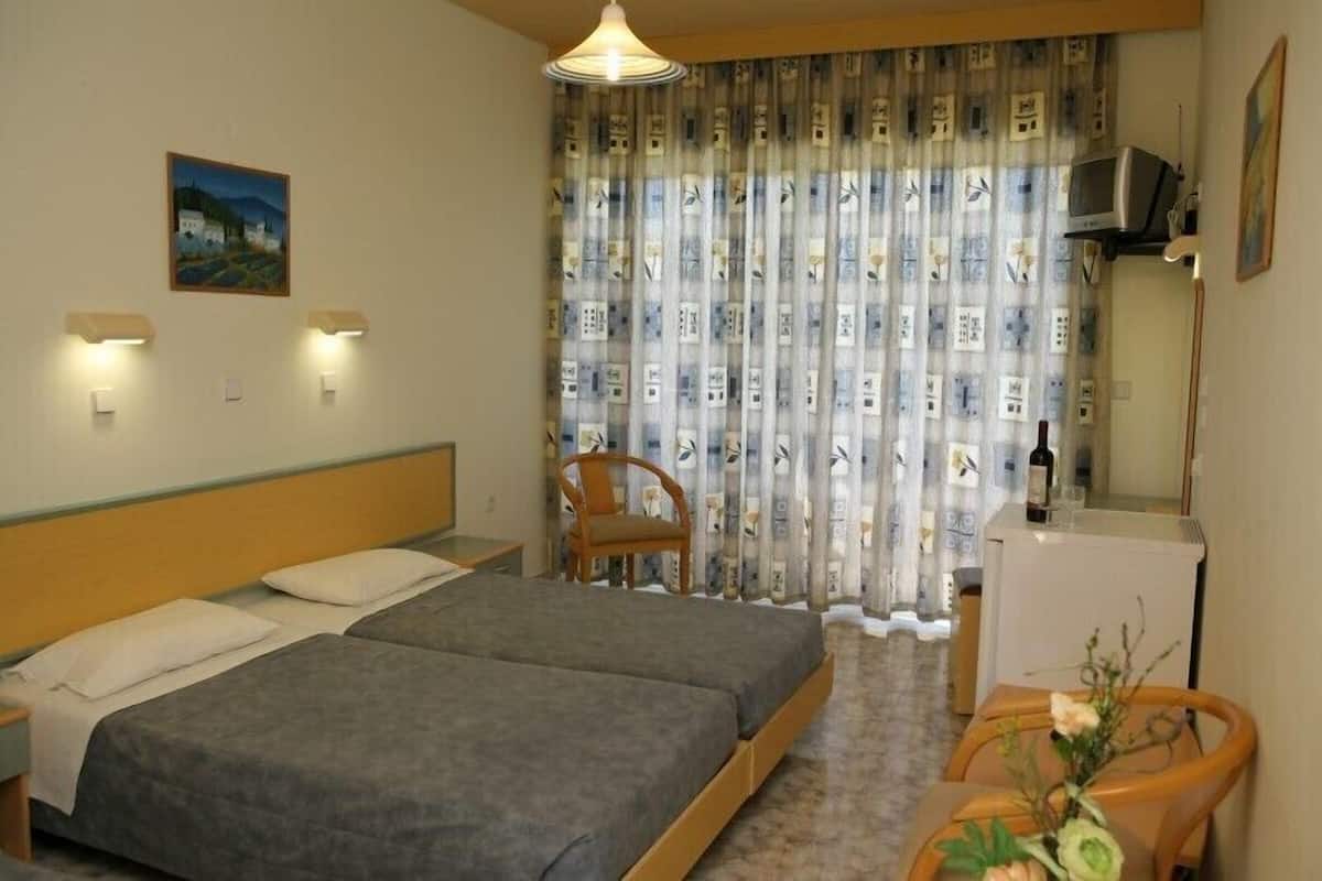 A hotel room with two single beds, a wooden headboard, a small table with a chair, a TV, and a refrigerator.