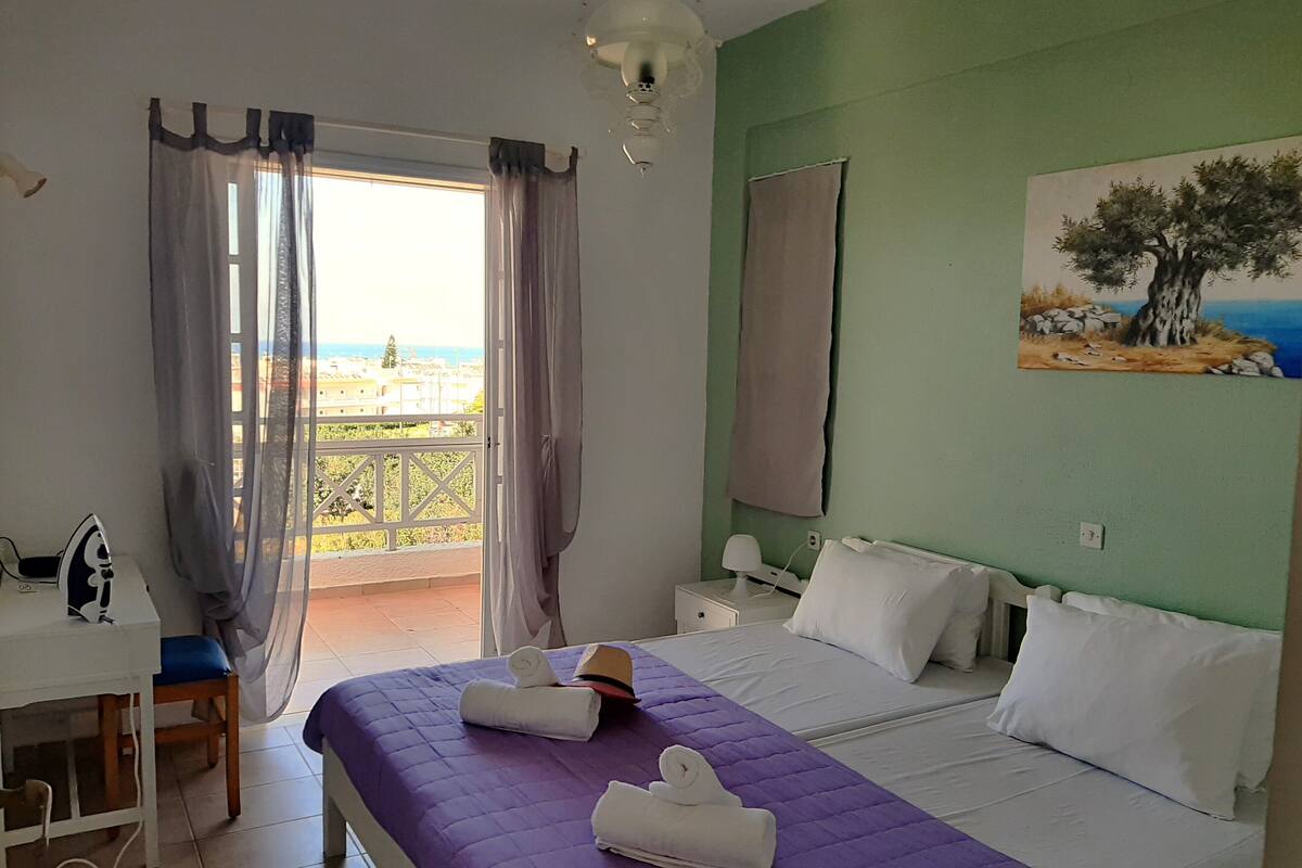 A hotel room with a bed, a desk, a chair, a painting, and a view of the sea.
