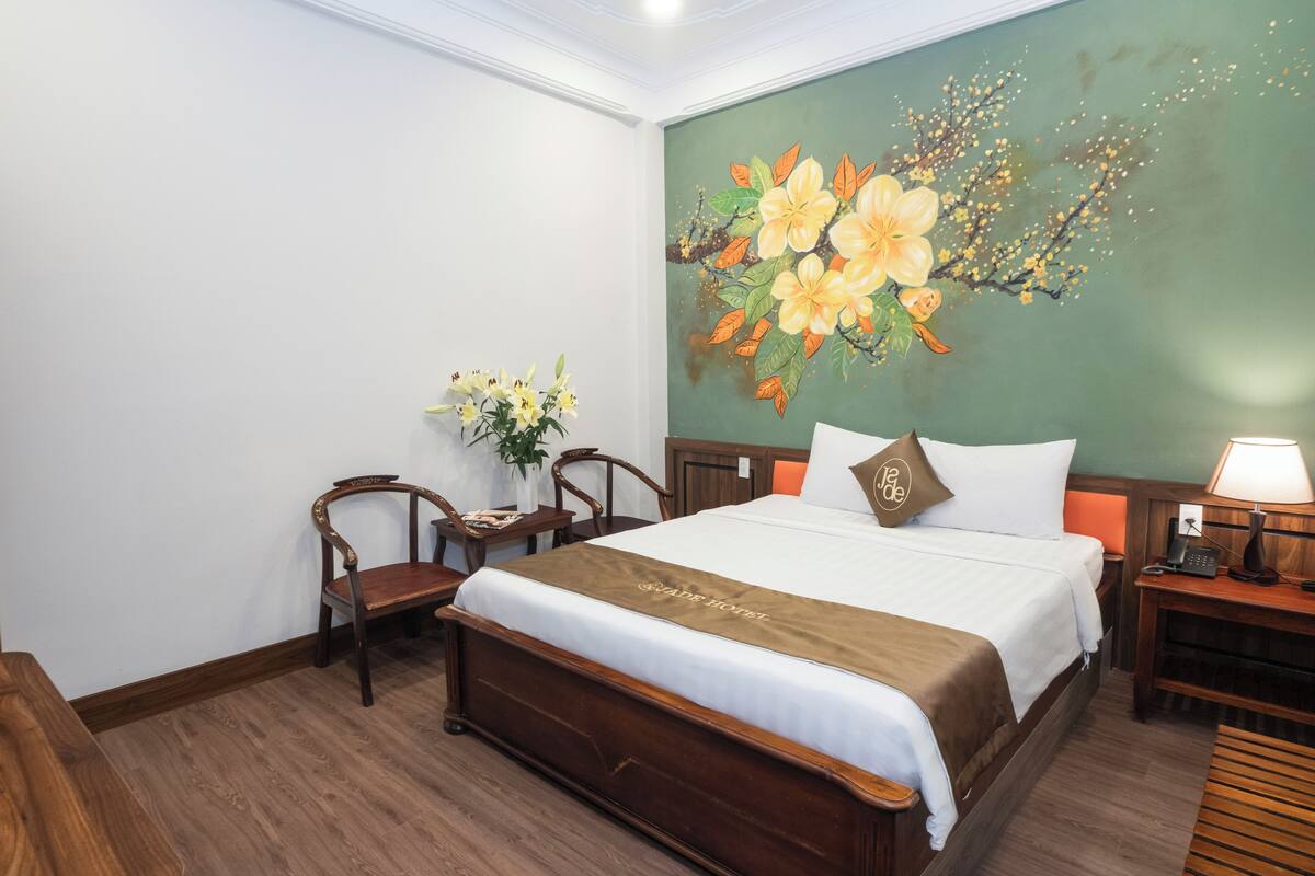 A hotel room with a large bed, wooden headboard, a nightstand with a lamp, a small table with a vase of flowers, and a wall painting of flowers.