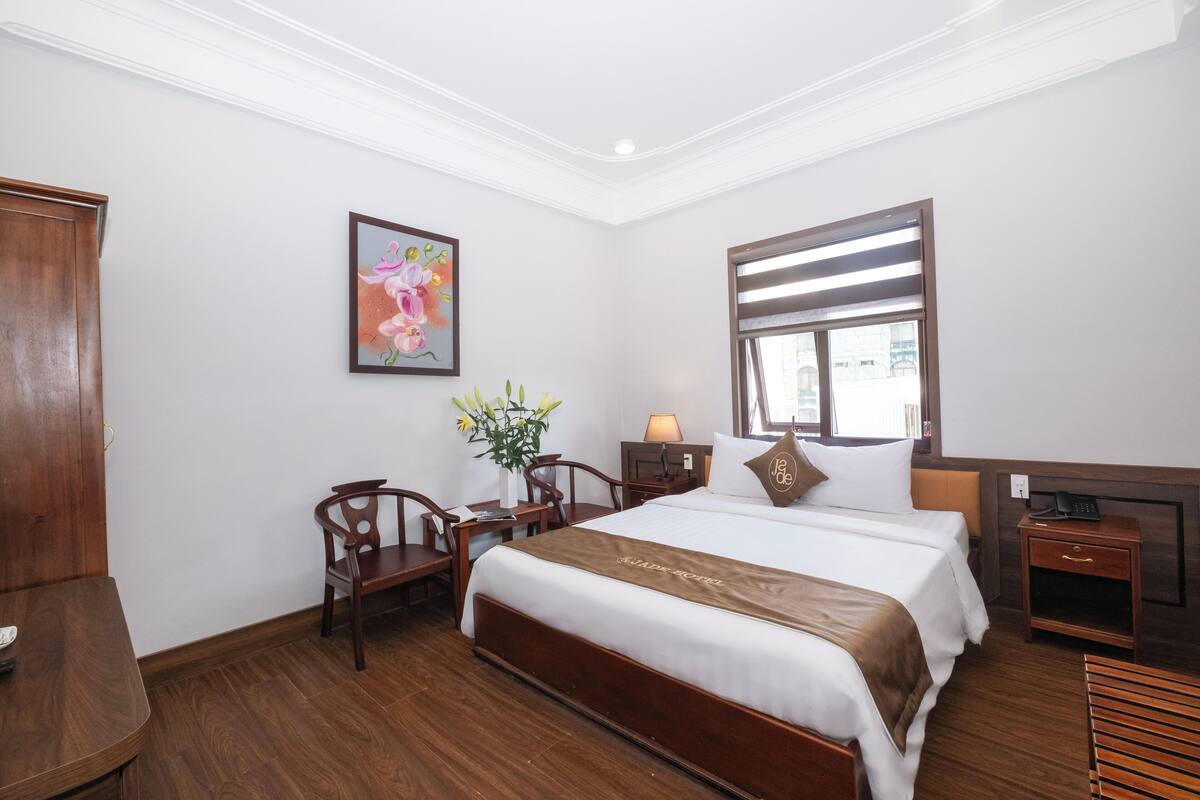 A hotel room with a large bed, a desk with a chair, a small table, a nightstand, a lamp, a framed picture on the wall, and a window with blinds.
