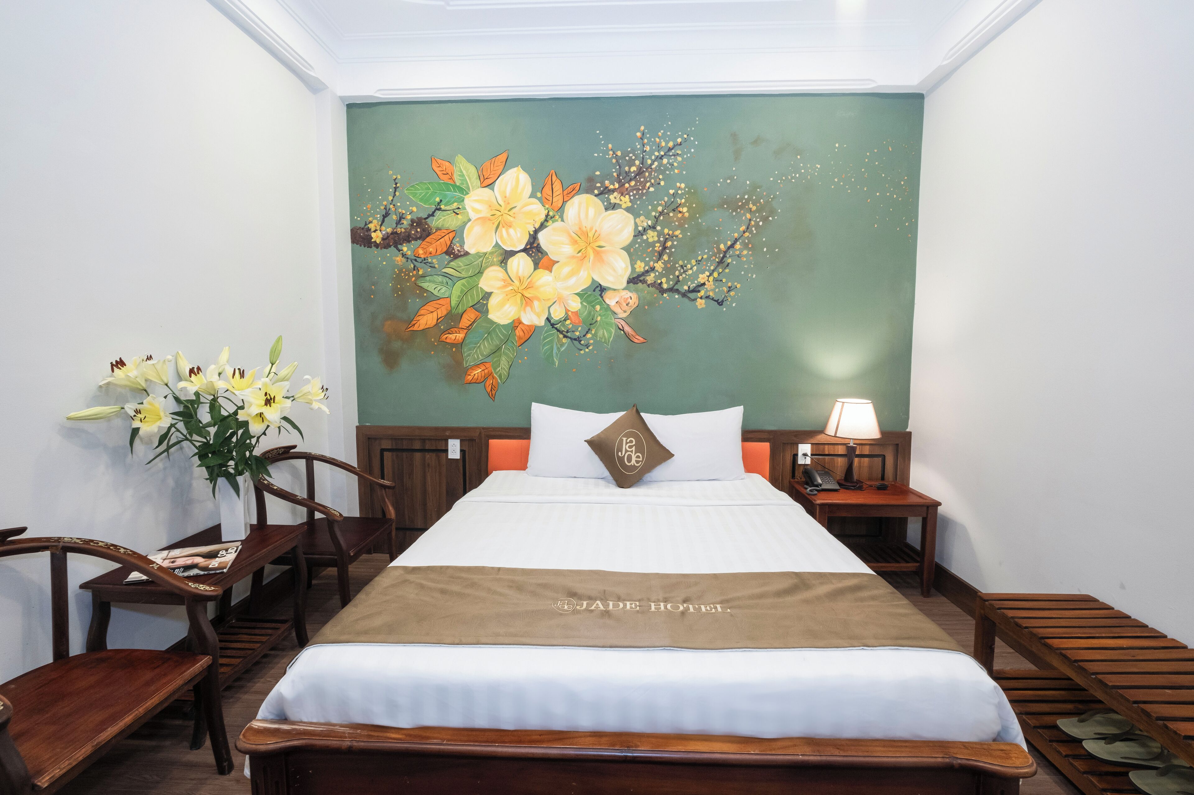 Habitación de hotel con cama, mesita de noche, lámpara, banco de madera y un mural floral en la pared.
