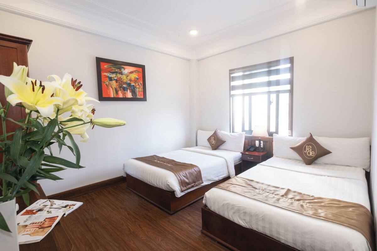 A hotel room with two beds, a large window, a painting on the wall, and a plant with yellow flowers.