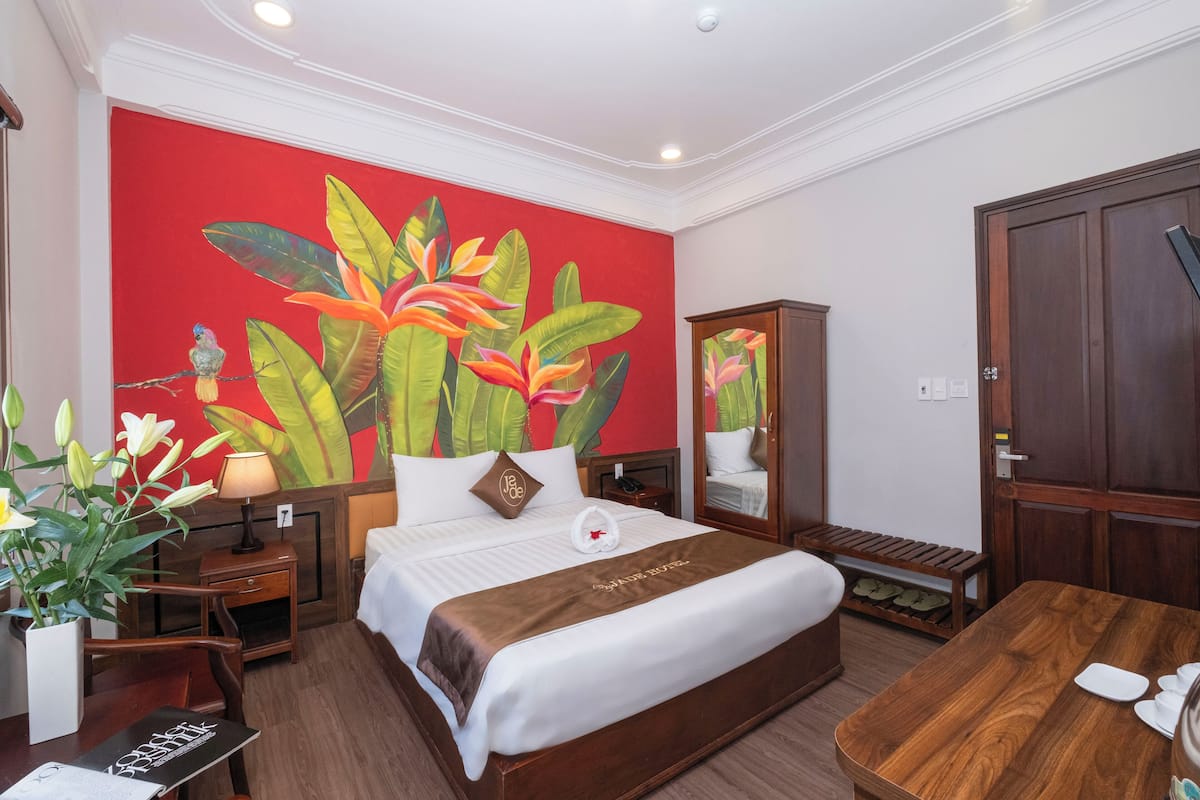 A hotel room with a large bed, a wooden desk, a nightstand, a mirror, and a red wall with a tropical mural.