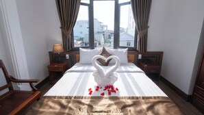 Habitación de hotel con una cama, una silla de madera, una mesita de noche, una lámpara y vistas a edificios a través de amplias ventanas.
