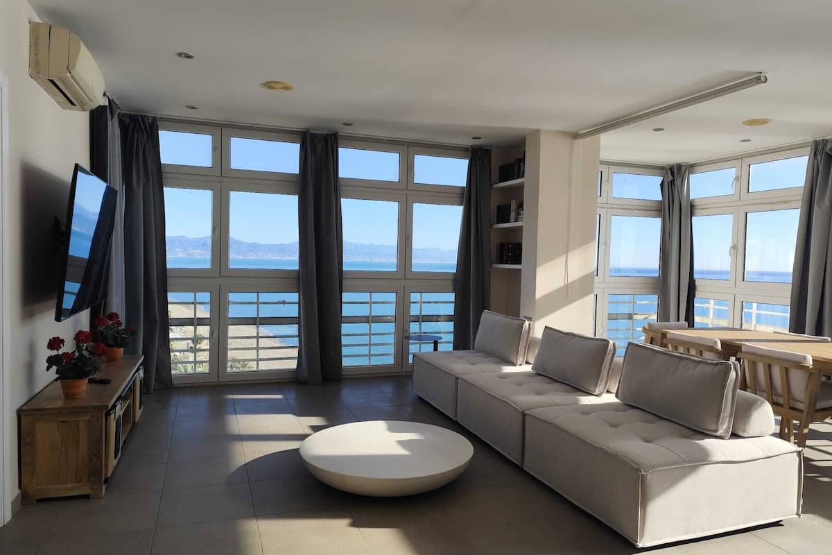 Una sala de estar moderna con un sofá grande, una mesa de centro redonda y un televisor sobre un mueble. La habitación tiene ventanas amplias con vistas al mar y a las montañas.