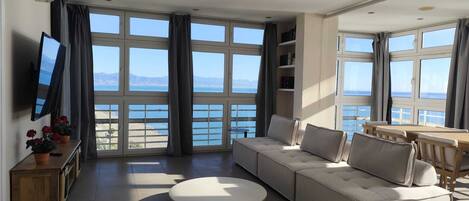 A modern living room with a large sofa, a round coffee table, and a TV on a stand. The room has large windows with a view of the sea and mountains.