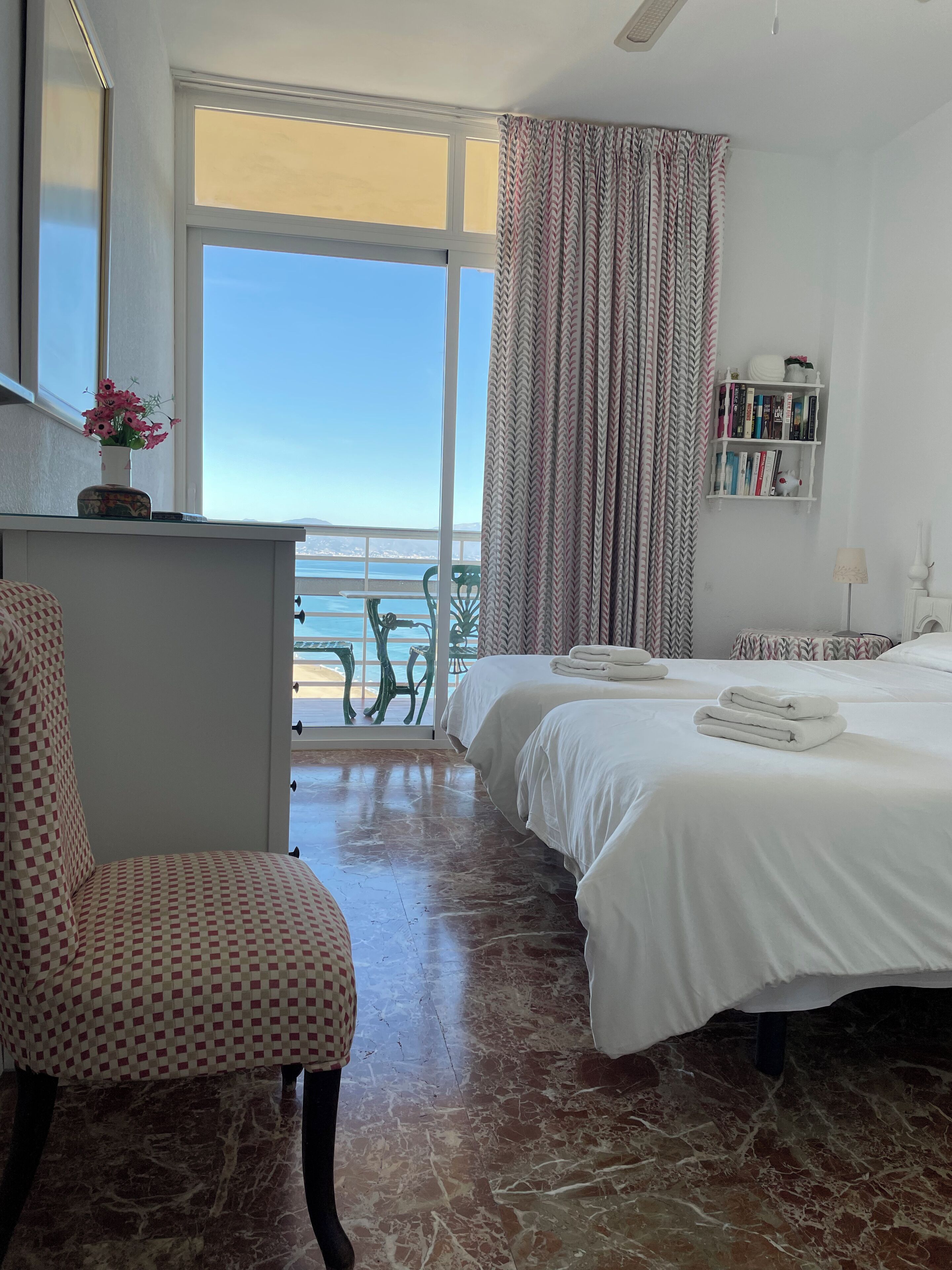 Une chambre d’hôtel avec un lit, une chaise, une petite table, une bibliothèque et une vue sur la mer.