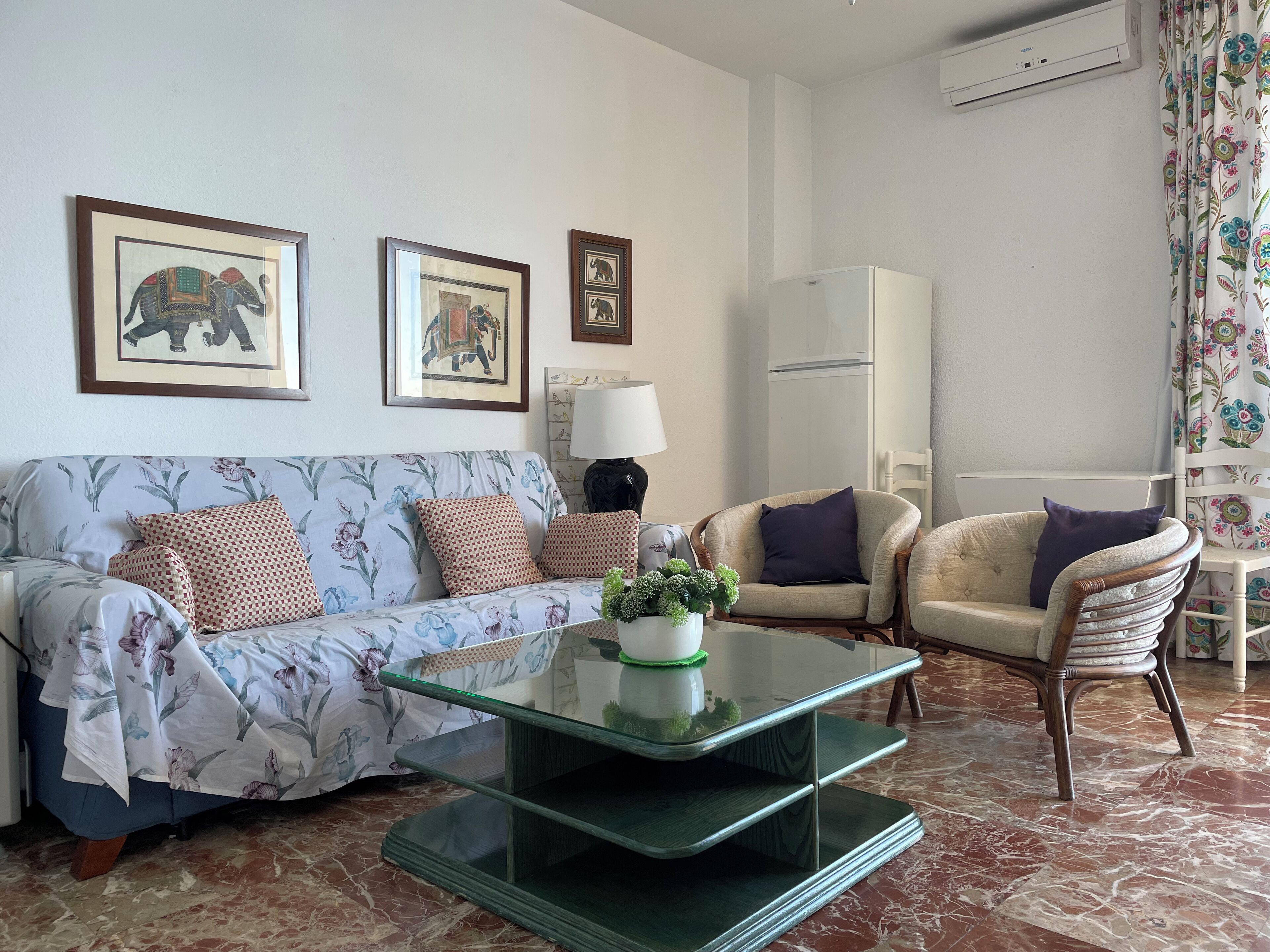 Una sala de estar con un sofá, sillones, una mesa de centro de vidrio y una mesita de noche con una lámpara.