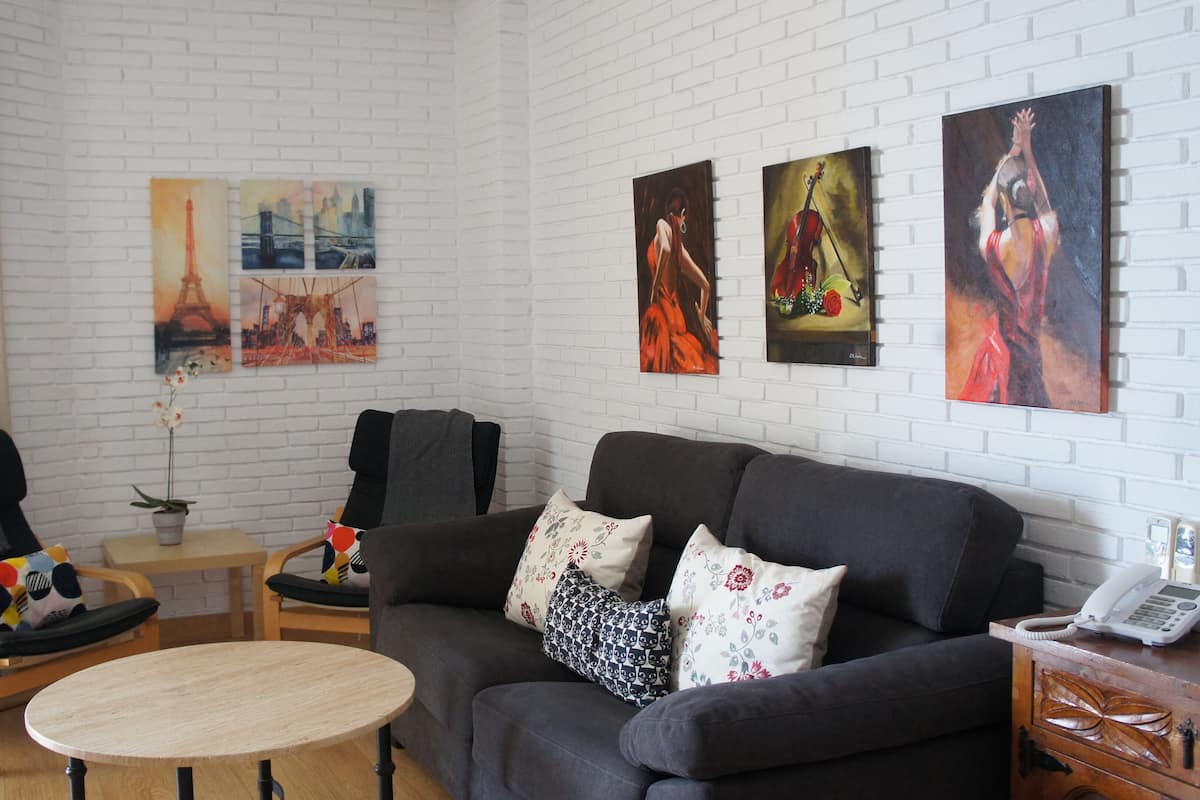 Una sala de estar con un sofá gris, dos sillones, una mesita de madera y una planta en maceta. Hay tres cuadros en la pared y un teléfono sobre una mesita auxiliar de madera.