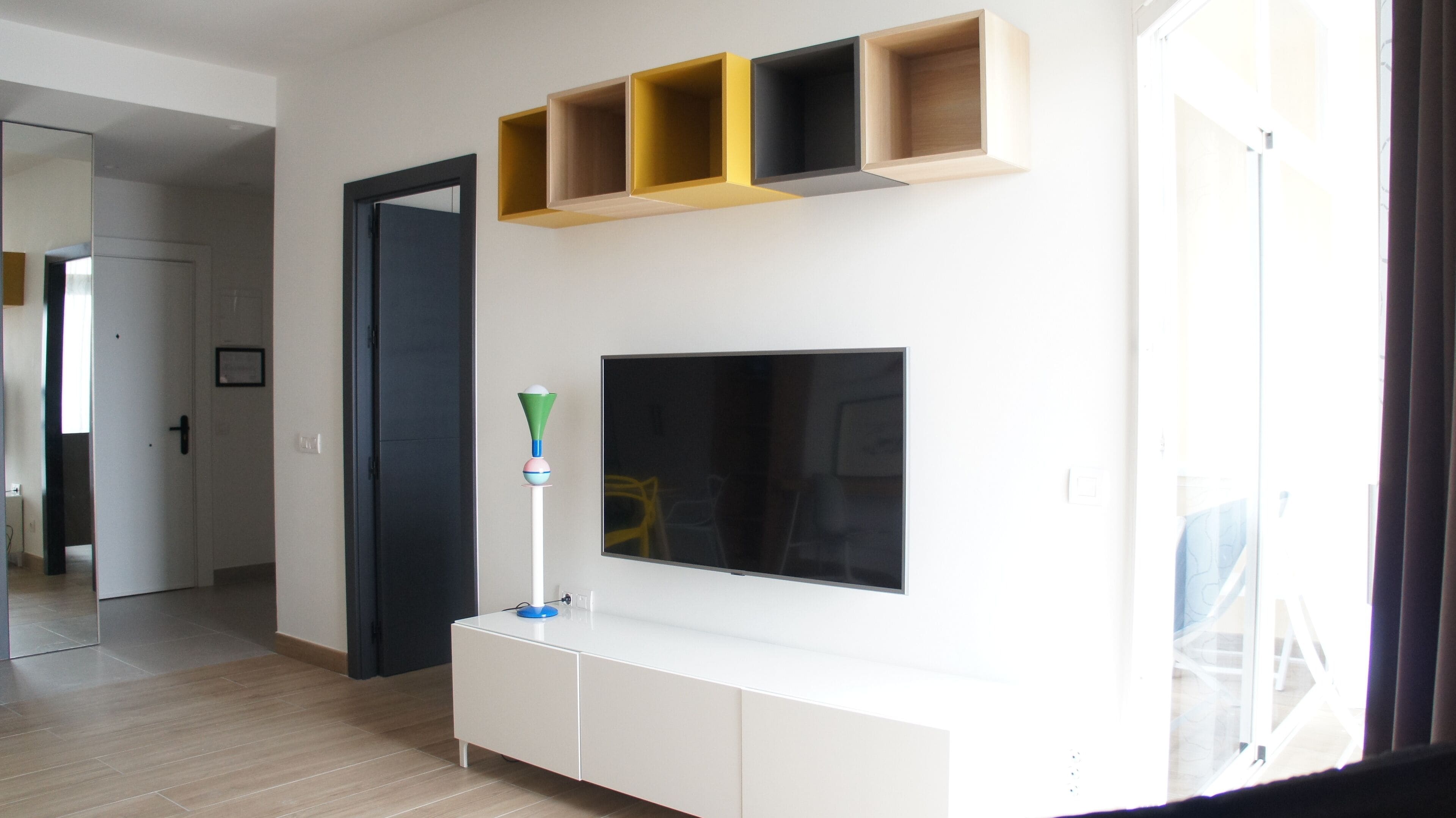 Un salon moderne avec un téléviseur à écran plat fixé sur une console blanche, des étagères en bois au-dessus, et une porte donnant sur une autre pièce.