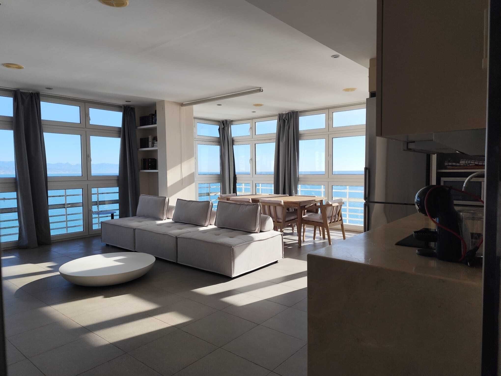 A modern living room with a sofa, a round coffee table, and a dining area with a table and chairs. Large windows offer a view of the sea.