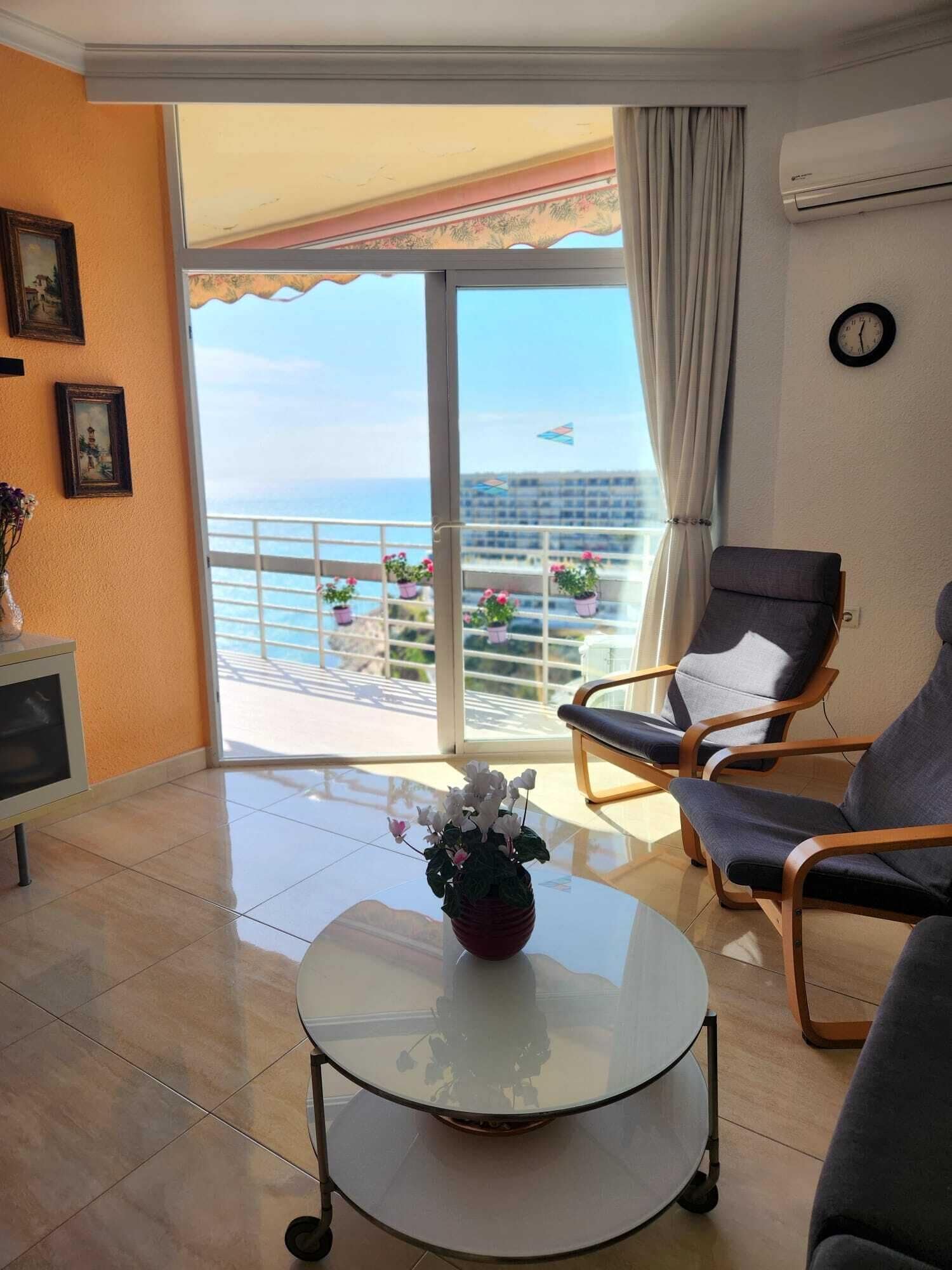 A room with a glass table, two armchairs, a small cabinet, and a view of the sea through a sliding door.