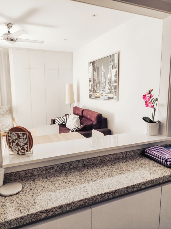 Un salon moderne avec un canapé blanc, une table basse, un tableau encadré représentant un paysage urbain, une orchidée en pot et un ventilateur de plafond.