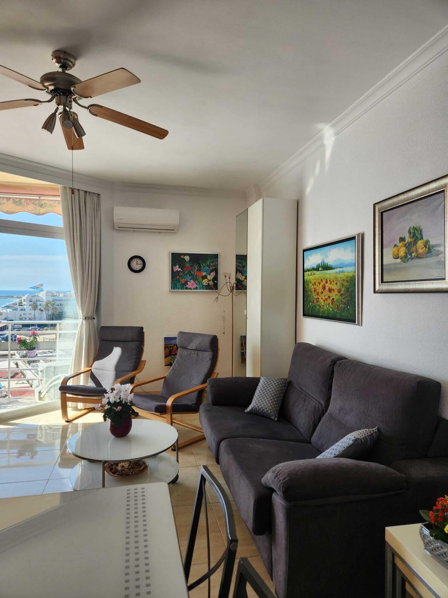 A modern living room with a ceiling fan, two armchairs, a sofa, and a coffee table. There are paintings on the wall and a view of the city through large windows.