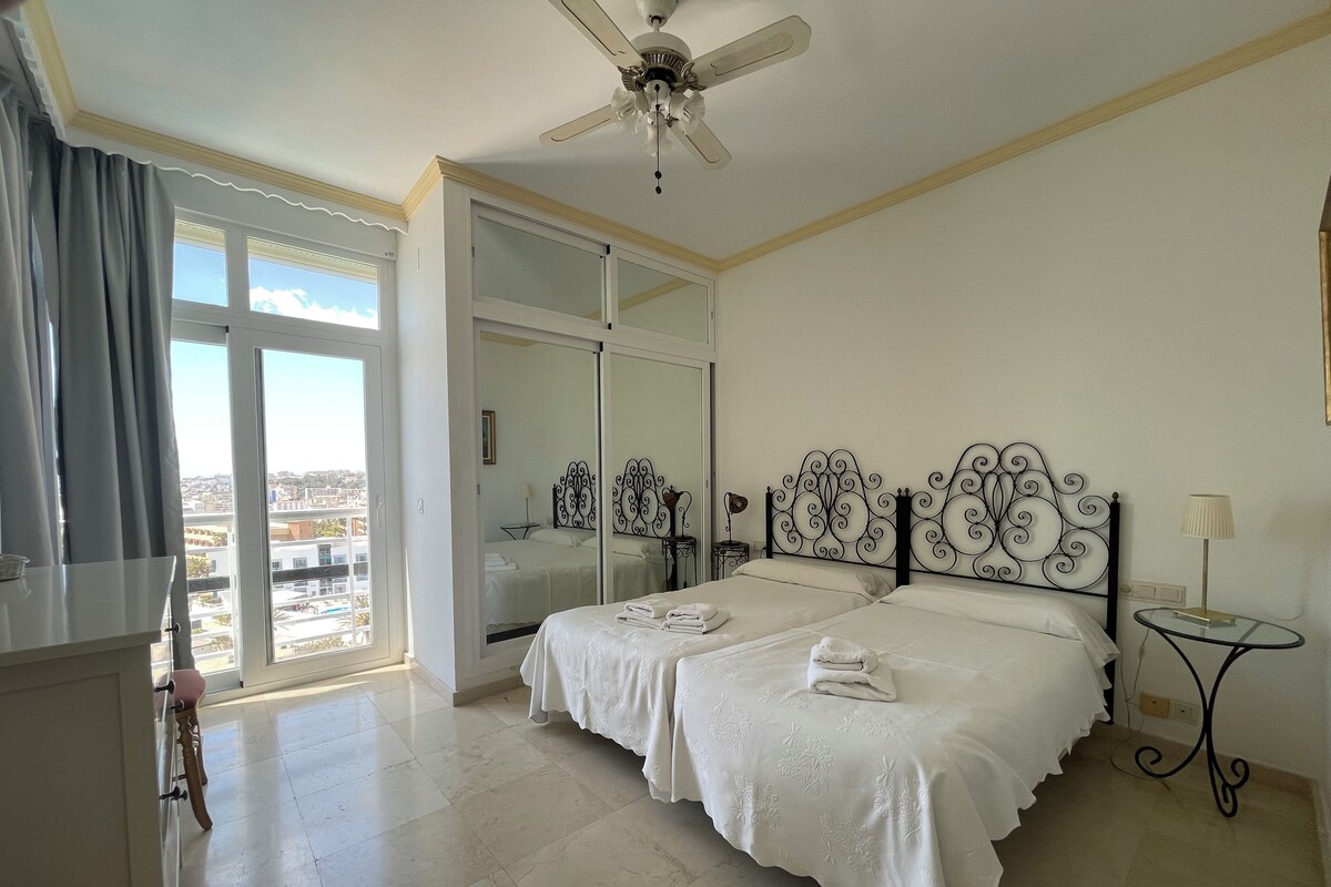 Una habitación de hotel con una cama, un ventilador de techo, una ventana con cortinas y vistas a la ciudad.