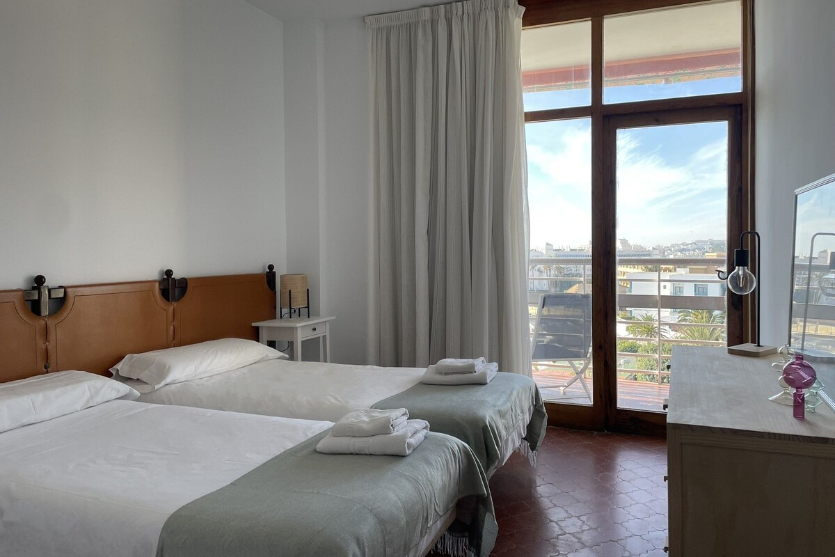 Habitación de hotel con dos camas, una ventana con cortinas, una mesita y vistas a la ciudad.