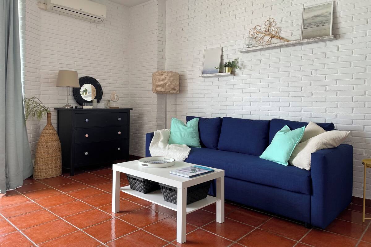 Una sala de estar con sofá azul, mesa de centro blanca, cómoda negra y pared de ladrillo blanco.
