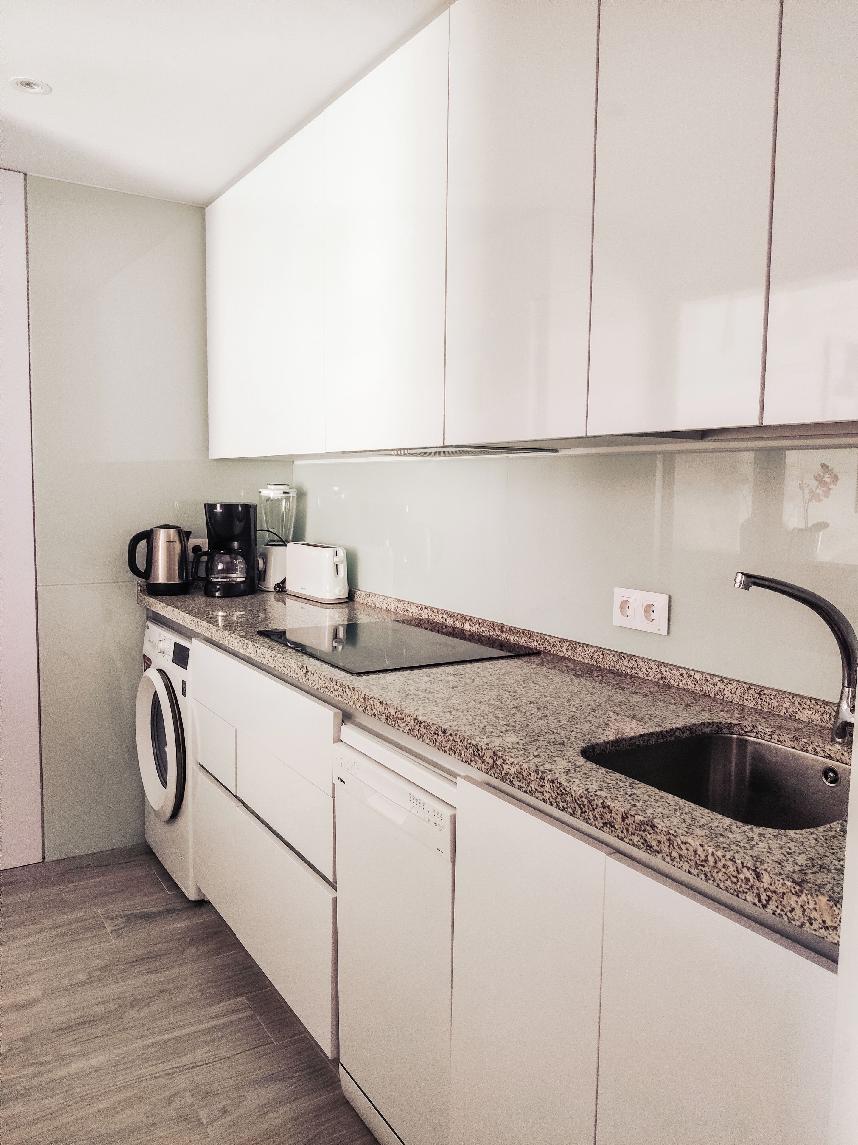 A modern kitchen with white cabinets, a granite countertop, a built-in dishwasher, a washing machine, and a coffee maker.