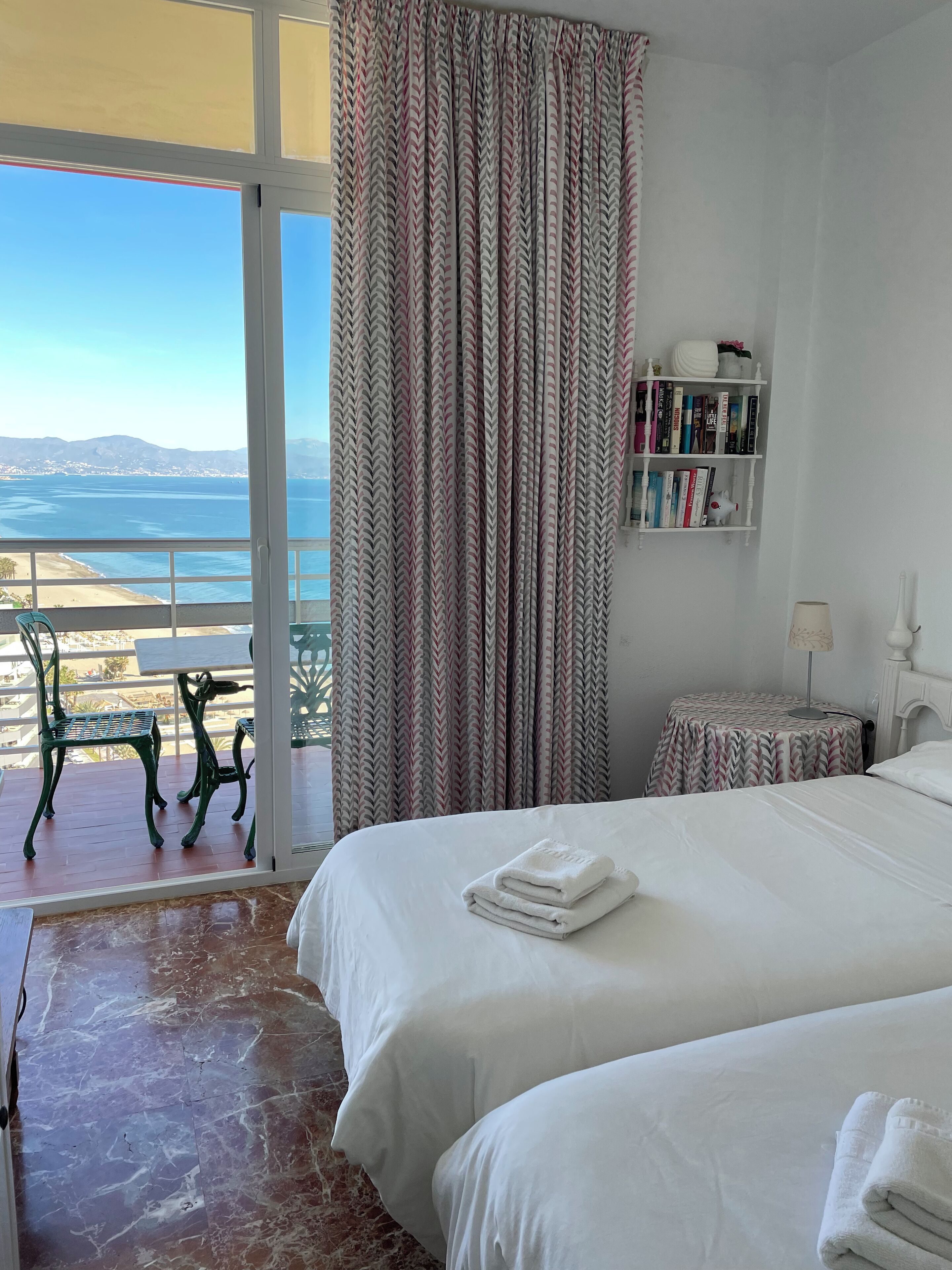 A hotel room with a large bed, a small table, a bookshelf, and a view of the beach and ocean.