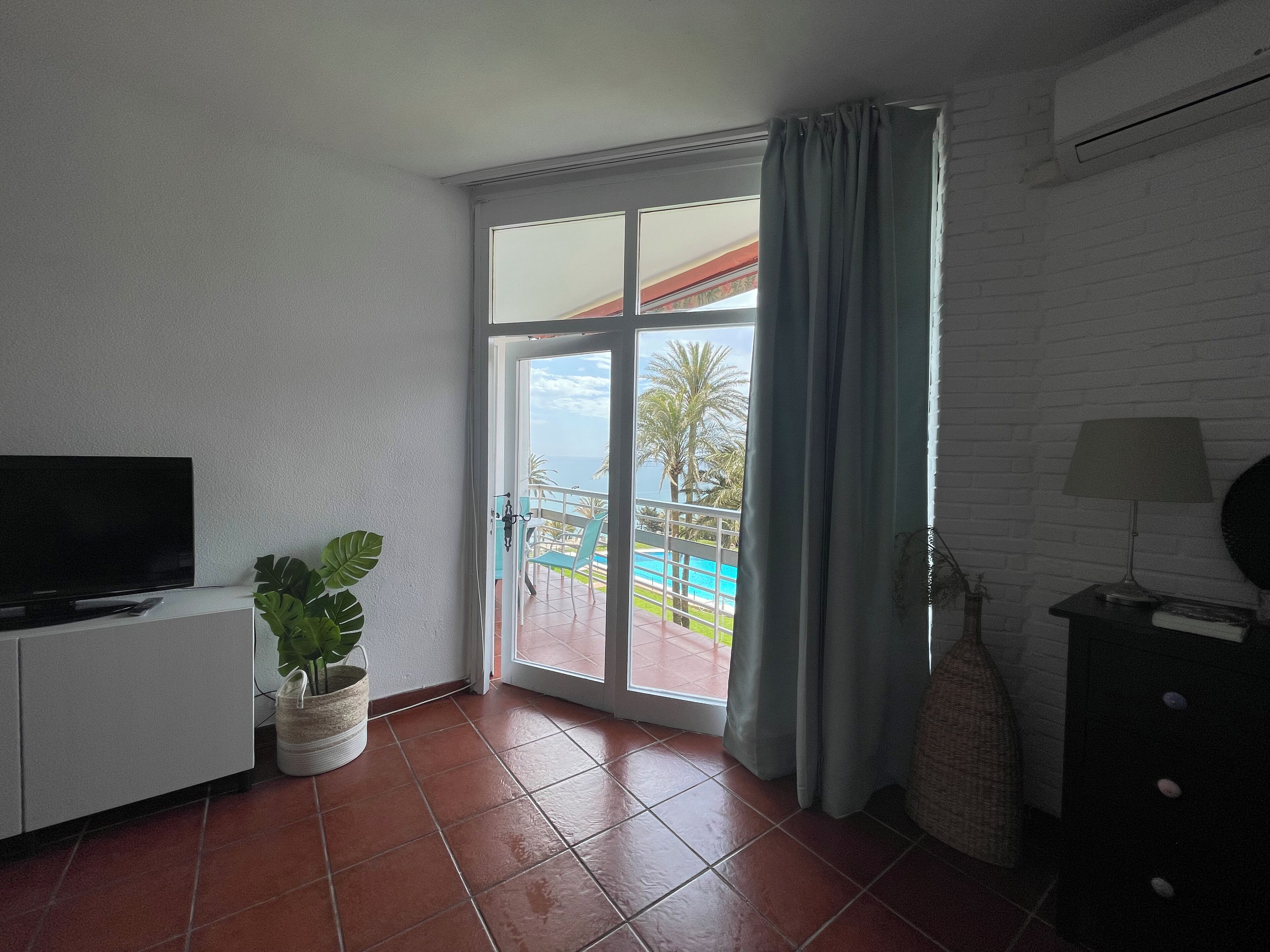 A room with a TV, a potted plant, a view of a swimming pool and palm trees, and a balcony with a railing.