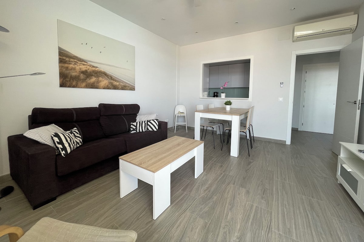 Una sala de estar moderna con un sofá marrón oscuro, una mesa de centro blanca y un comedor con una mesa y sillas blancas.