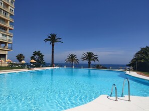 Une piscine d’un bleu éclatant, avec vue sur l’océan, entourée de palmiers et bordée d’un immeuble à plusieurs étages.