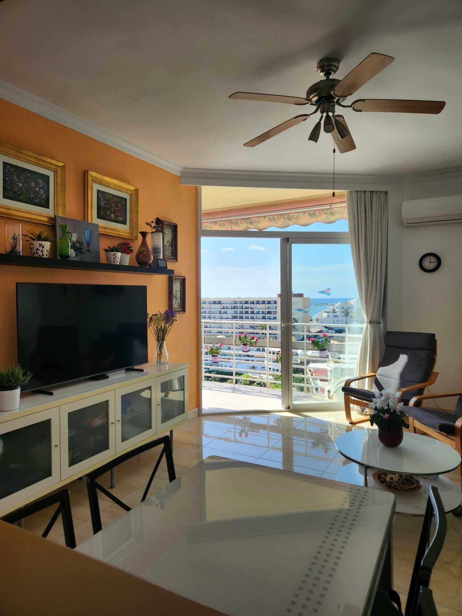 Una sala de estar con televisor, ventilador de techo, balcón con vistas a la ciudad y comedor.