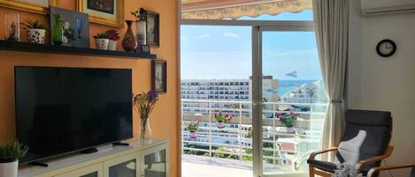 Una sala de estar con televisor, ventilador de techo, balcón con vistas a la ciudad y comedor.