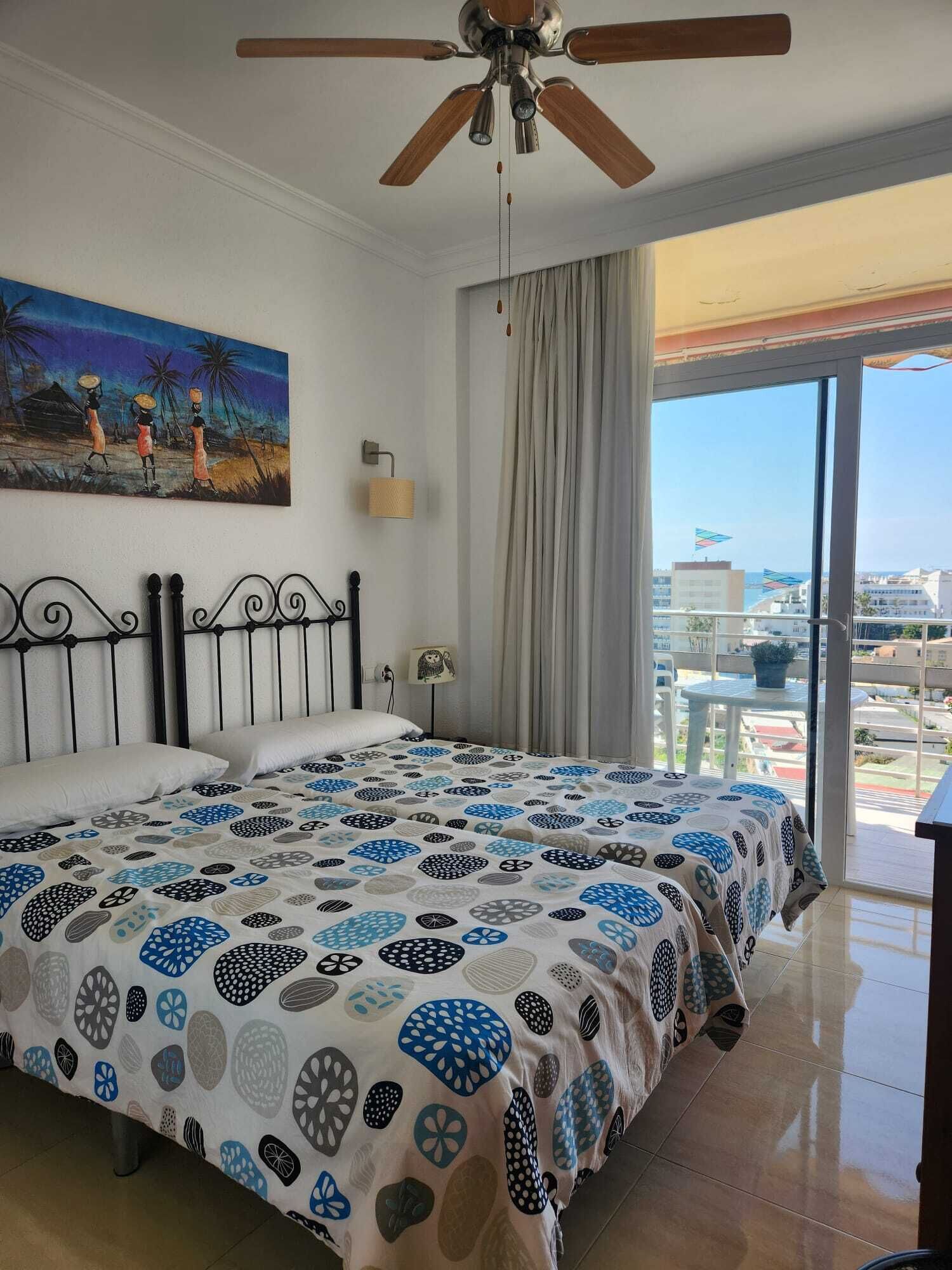 Habitación de hotel con cama, ventilador de techo, un cuadro, una ventana con cortinas y vista a la ciudad.