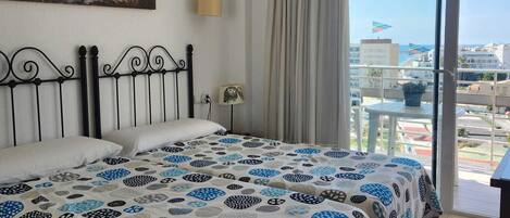 Habitación de hotel con cama, ventilador de techo, un cuadro, una ventana con cortinas y vista a la ciudad.