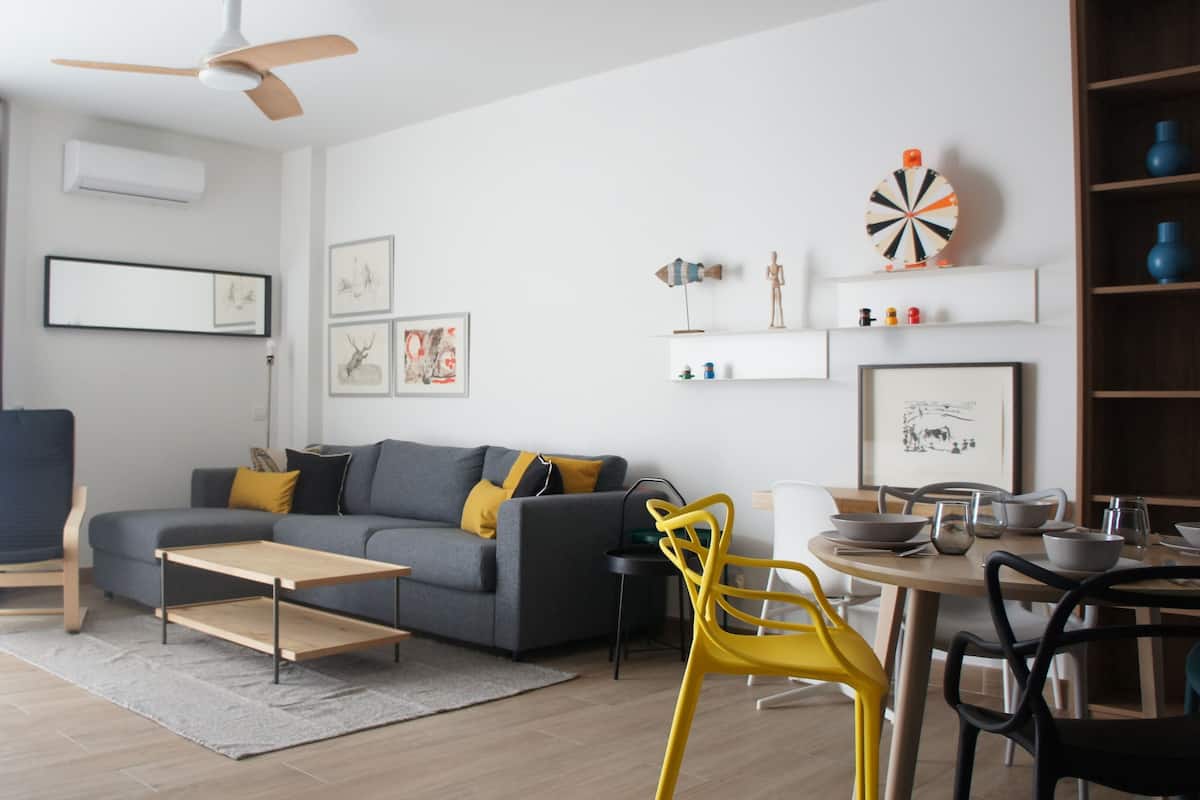 Una sala moderna con un sofá gris, cojines amarillos, una mesa de centro de madera y un comedor con una silla amarilla y una mesa preparada para dos comensales.