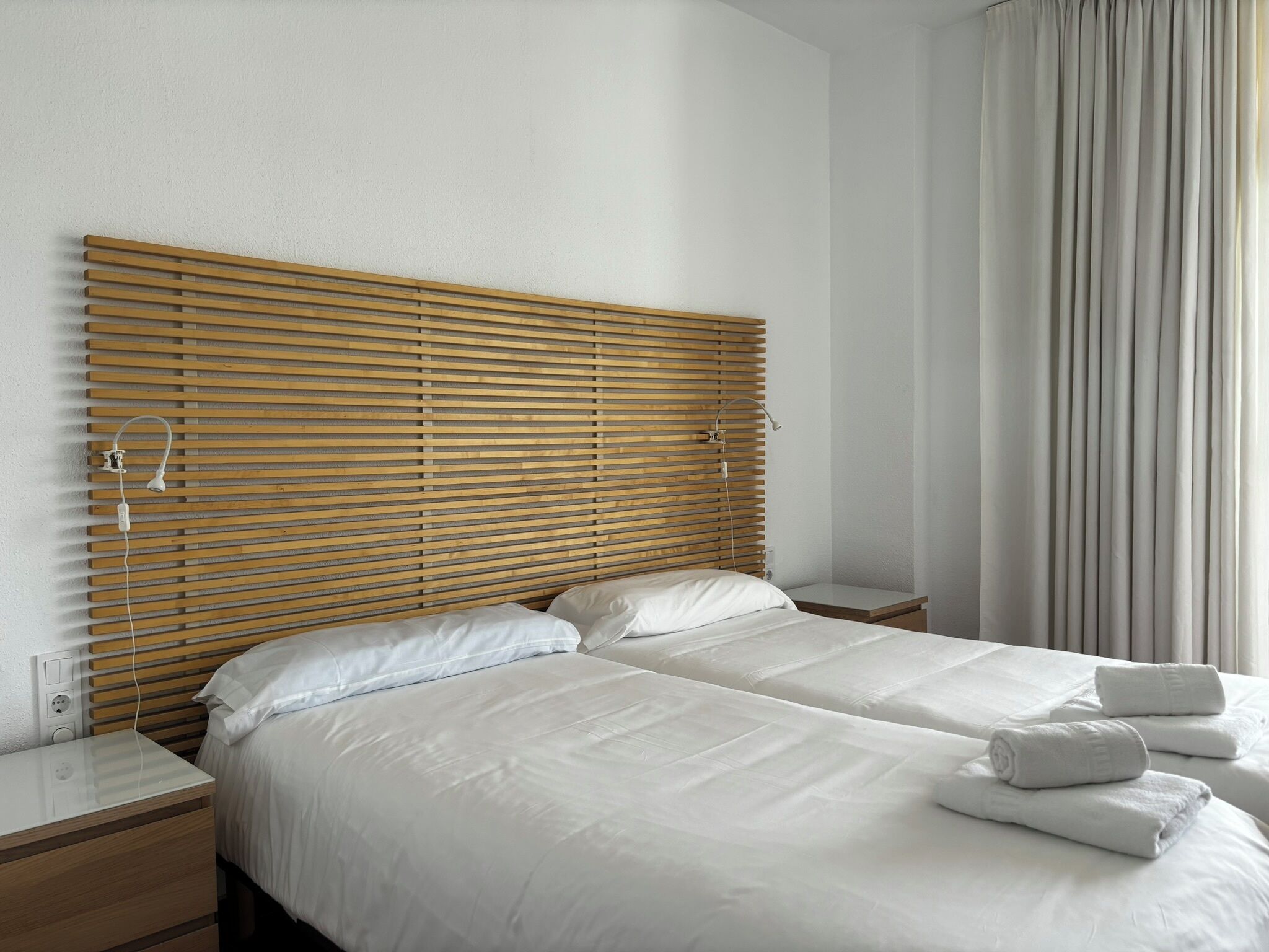 A hotel room with a wooden slatted headboard, two beds with white linens, a bedside table, and a window with curtains.