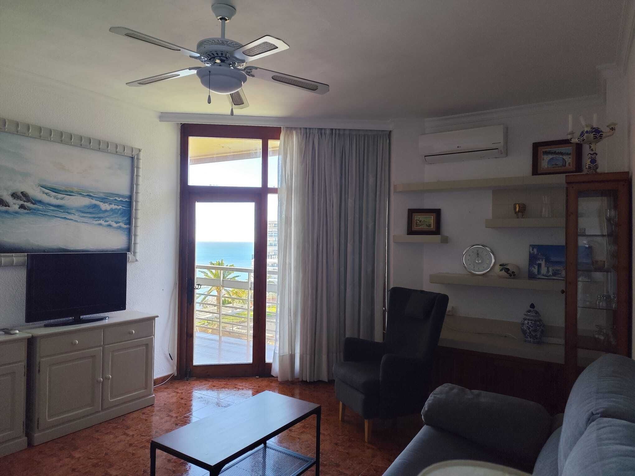 Uma sala de estar com televisão, ventilador de teto, uma cadeira, um sofá e vista para o oceano através de uma grande janela.