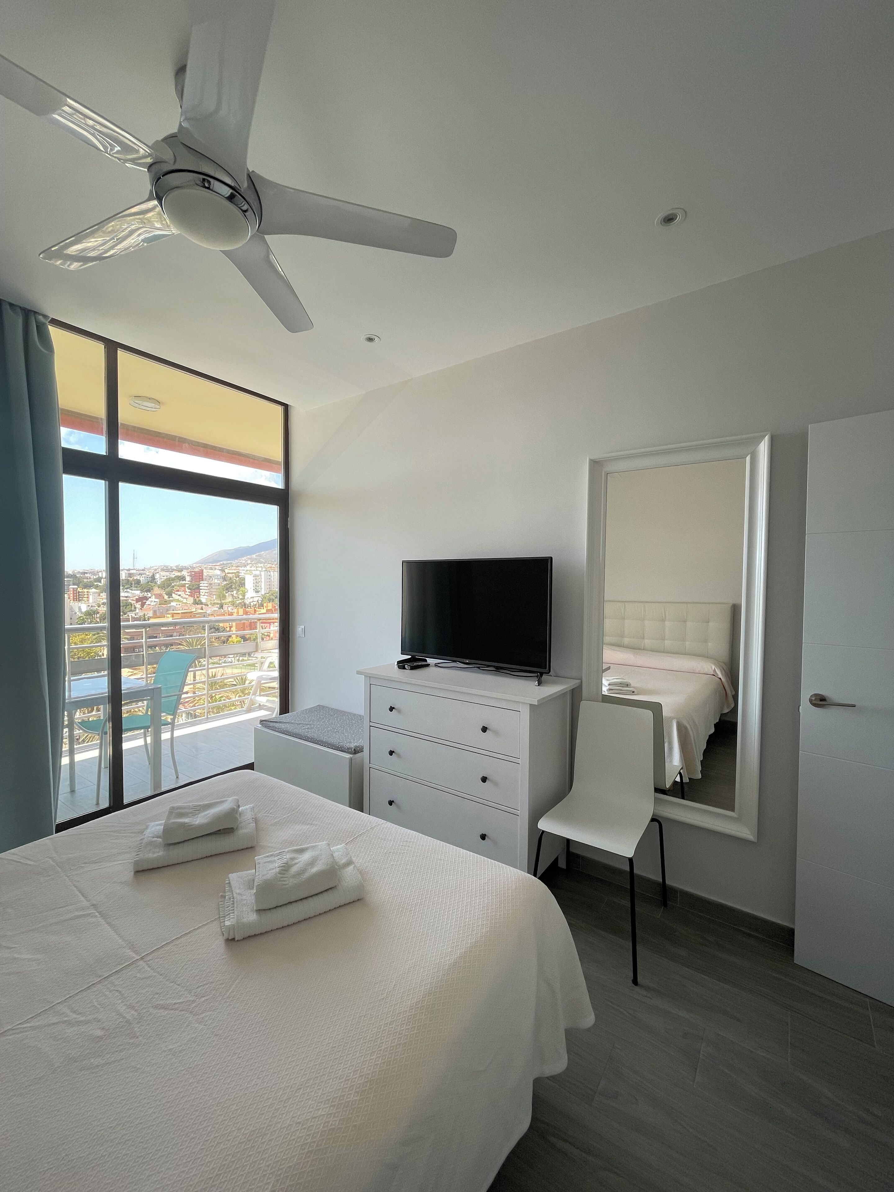 Une chambre d’hôtel comprenant un lit, un bureau avec une télévision, une chaise, une commode, un ventilateur de plafond et une grande fenêtre donnant sur la ville.