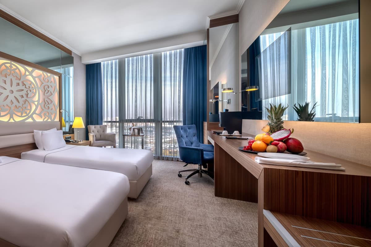 A hotel room with two beds, a desk, a chair, and a large window with curtains.