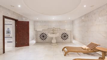 A spa room with a marble floor, wooden loungers, a central fountain, and decorative wall panels.