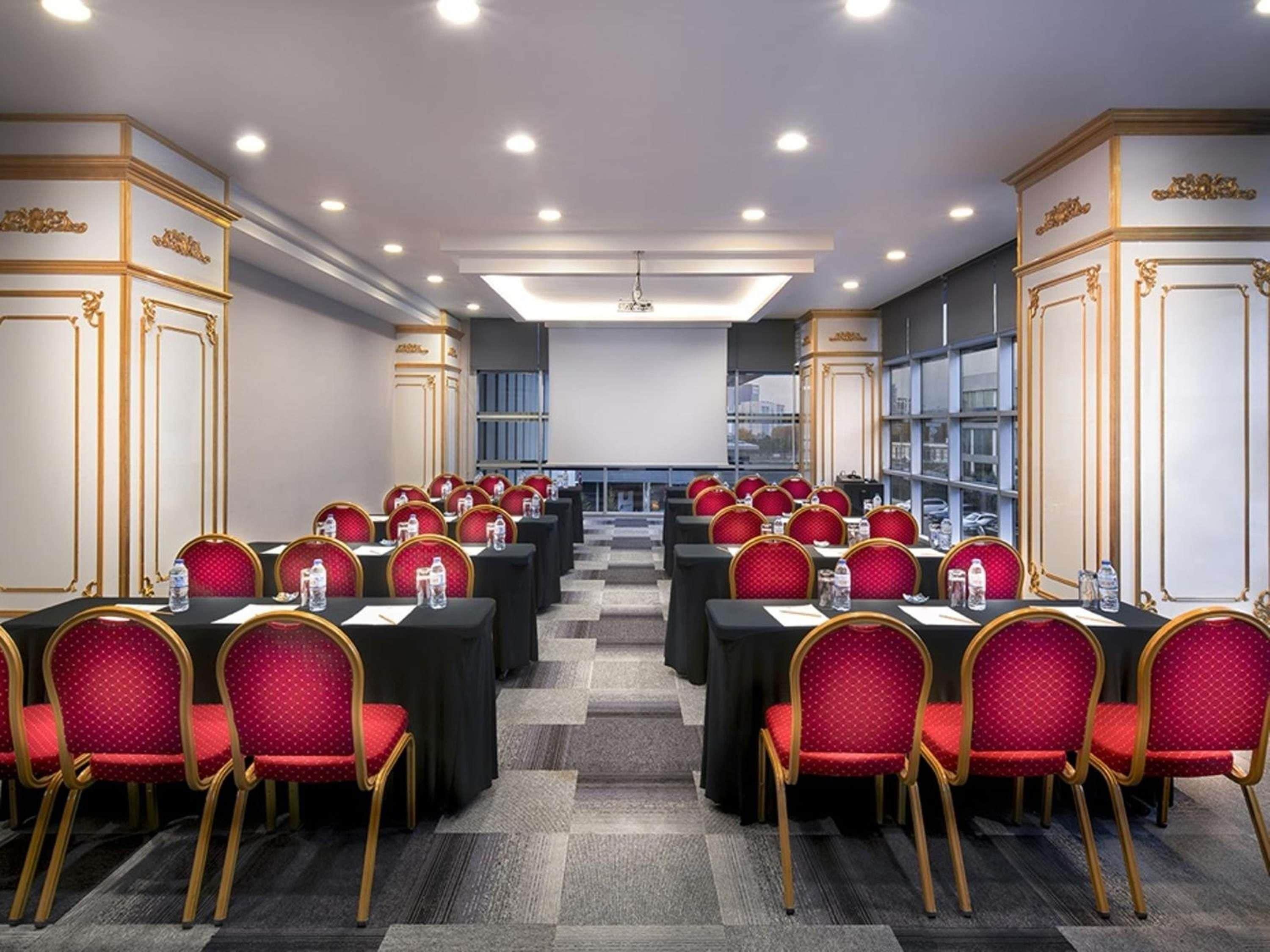 A conference room with rows of tables and chairs, equipped with water bottles, and a large screen at the front.